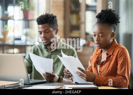Two young African colleagues in casualwear looking through papers while sitting by table next to one another Stock Photo