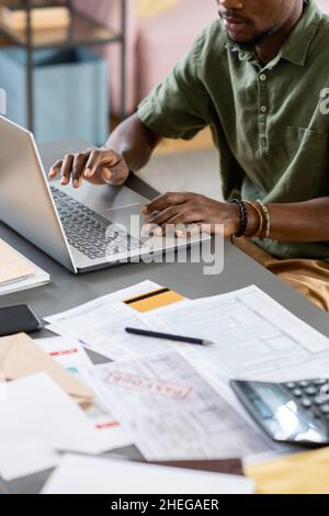 Hands of young African male economist working in front of laptop while sitting by table with financial documents Stock Photo