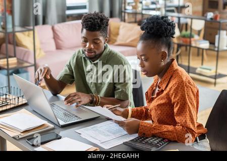 Young confident businessman making presentation to African female with papers while checking online data and that in documents Stock Photo