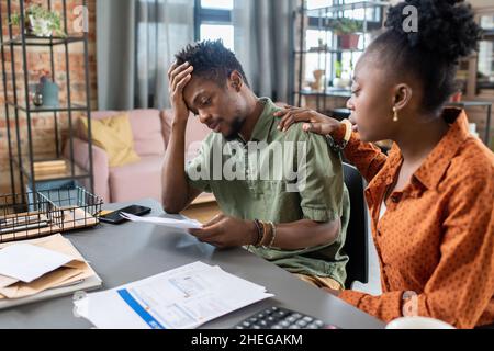 Young African female keeping hand on shoulder of stressed male colleague with financial papers while comforting him Stock Photo