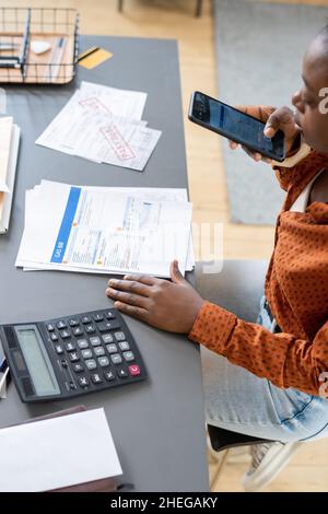 Young businesswoman with smartphone taking photo of documents while sitting by table with calculator, papers and office supplies Stock Photo