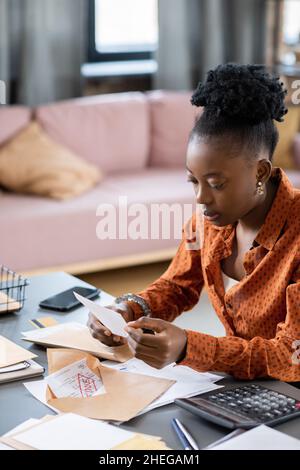 Young African female looking through financial paper or receit while sitting by table with documents and unsealed envelope Stock Photo