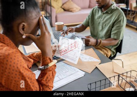 Young stressed female looking at unpaid bill and touching her head while sitting by table in front of man networking Stock Photo