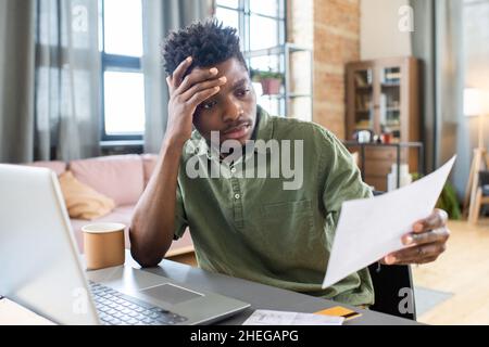 Young stressed African businessman looking through unpaid bill and touching his head while sitting in front of laptop Stock Photo