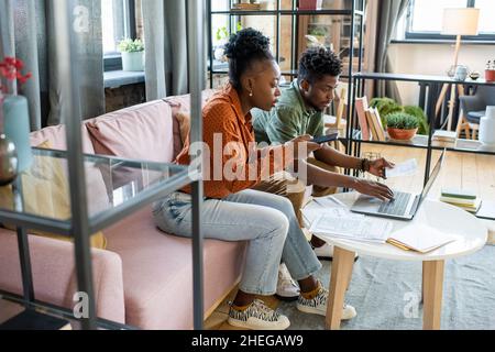 Two young contemporary coworkers working with mobile gadgets and financial data while sitting on couch by table Stock Photo