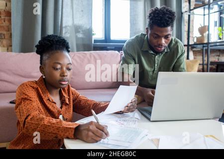 Two young African colleagues working with financial papers and computer data while sitting by white round table Stock Photo