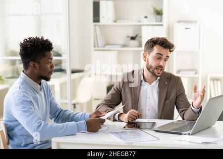 Confident businessman making presentation of online data on laptop display to young African colleague at working meeting in office Stock Photo