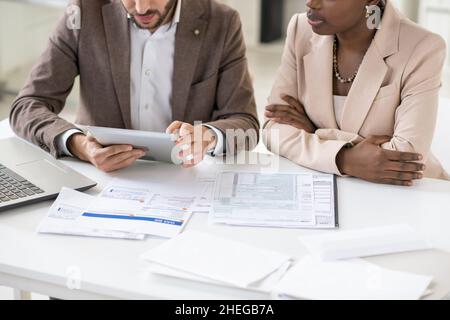 Two young contemporary intercultural colleagues in formalwear discussing data in tablet while sitting by table with financial documents Stock Photo