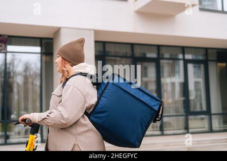 Side view of female courier food delivery with big thermal backpack riding on electric scooter on city street. Stock Photo