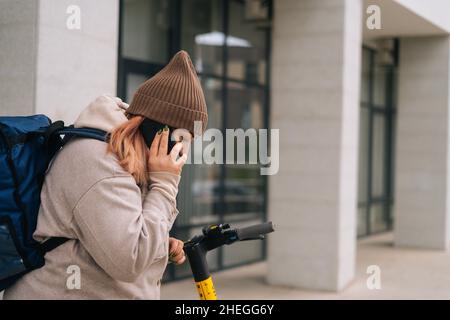 Side view of female courier food delivery in warm clothes calling to client on mobile phone standing at street with electric scooter. Stock Photo