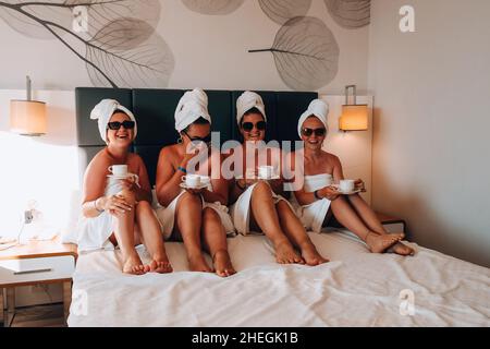 Four girls friends in white towels sitting on the bed having fun in hotel. Vacation Bloggers sunglasses drinking coffee together. Chat sweetly, tell stories and laugh. Friends met and spend their leisure time in hotel room. Concept of friendship Stock Photo