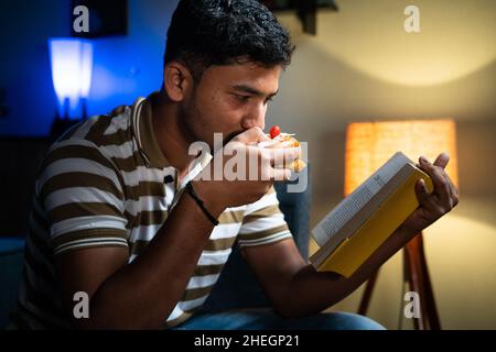 Young indian student eating unhealthy food while reading book at home - concept of unhealthy lifestyle, hobby, skill and knowladge development Stock Photo