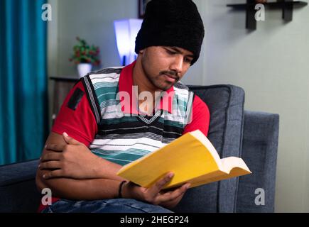 Young man reading books by wearing sweater and cap during freezing winter season at home - concept of hard and smart work, studying for exam, skill Stock Photo