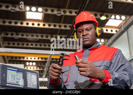 Portrait Of Black Machine Operator In Red Helmet, Hearing Protectors And Work Uniform Next To CNC Controller. Stock Photo
