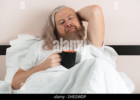 Bearded man lying in cozy bed after waking up holding cup of coffee with his eyes closed enjoying and dreaming being in harmony with himself Stock Photo