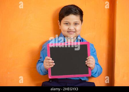 Portrait of happy cute little indian boy in school uniform holding blank slate against orange background, Adorable elementary kid showing black board. Stock Photo
