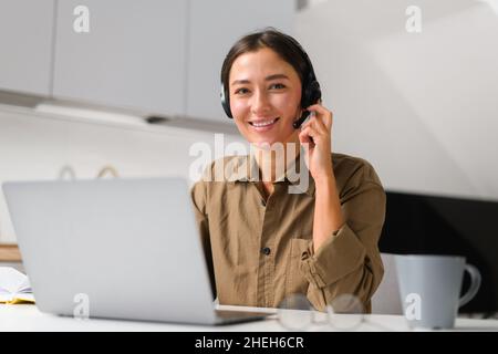 Smiling asian female freelancer wearing headset with microphone using laptop for online connection with colleagues or customers working remotely, woman sitting in the home office ad looks at camera Stock Photo