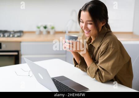 Relaxed asian multiracial female freelancer relaxing sitting with cup of coffee in front of laptop, working or studying remotely from home, drinking from mug taking a break Stock Photo