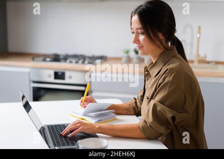 Female student watching a lecture, webinar using a laptop, sits at the table and writes with a pen in a notepad. Asian young woman employee takes a note watching online training, working remotely Stock Photo