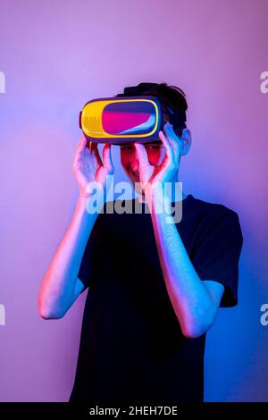 A young man looks out from under a virtual reality helmet and laughs, neon colors. Stock Photo