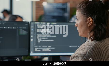 Over shoulder view of focused database designer writing code looking at multiple computer screens displaying algorithm. System engineer coding while colleagues doing teamwork in background. Stock Photo