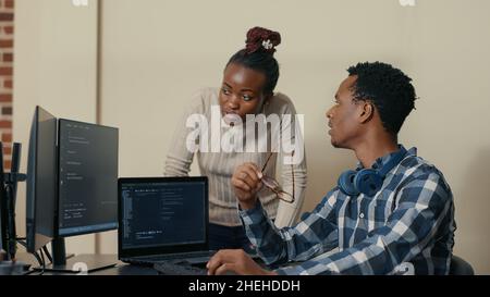 Database developer removing glasses while explaining algorithm to colleague coder pointing at running code on multiple computer screens. System engineers collaborating on development group project. Stock Photo