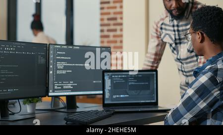 Focused software developer writing code on laptop looking at multiple screens with programming language is interrupted by colleague coder asking for advice. Programmers doing online cloud computing. Stock Photo