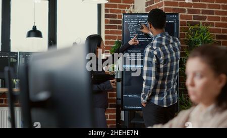 System developers analyzing code on wall screen tv looking for errors comparing results on digital tablet next to team of coders. Programmers working together in it startup company. Stock Photo