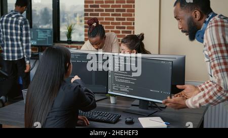 Mixed programers team working on group project on multiple screens showing running code in it startup office. Coder pointing pencil at computer screen with software compiling code. Stock Photo