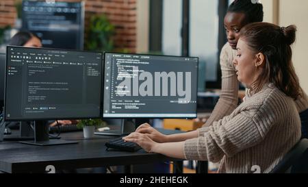 Junior programer writing algorithm under experienced colleague supervision sitting at desk with multiple screens showing programming interface. Developer writing code with the help of colleague. Stock Photo