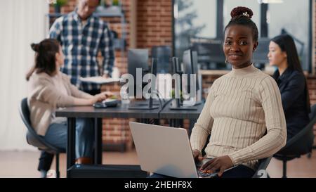 Portrait of african american coder sitting down writing programming language on laptop looking up and smiling at camera. System egineer using portable computer in software development agency. Stock Photo