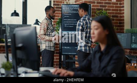 Programmers leaving after analyzing source code on wall screen tv comparing errors using digital tablet with focus switching to programer writing algorithm. Team of coders working on group project. Stock Photo