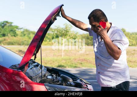 Man trying to call service, standing in front of the open hood of a broken down car outside highway Stock Photo