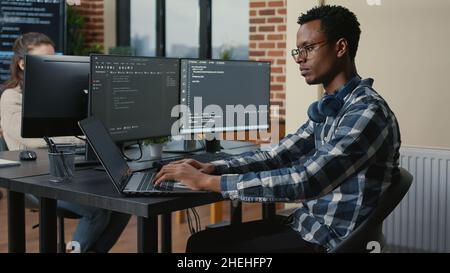 Programer thinking while touching beard and fixing glasses while typing on laptop sitting at desk with multiple screens parsing code. Focused database admin working with team coding in the background. Stock Photo