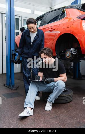 Young workman writing on clipboard on tire near colleague with screwdriver in car service Stock Photo
