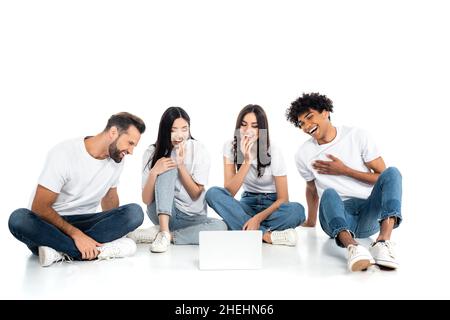 full length view of multiethnic friends laughing while watching comedy film on laptop on white Stock Photo