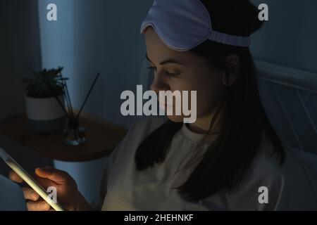Woman in silk mask using laptop computer and surfing social media on smartphone in bed before sleep at night. Social media addiction concept Stock Photo