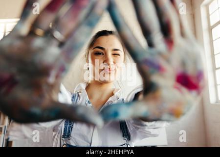 Carefree young painter framing her face with her hands. Happy female artist winking at the camera while standing in her art studio. Creative young wom Stock Photo