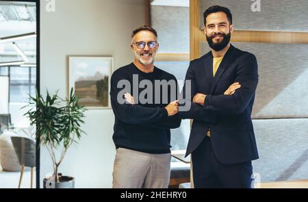 Two successful businessmen smiling at the camera while standing with their arms crossed. Two cheerful businessmen working together in a modern co-work Stock Photo