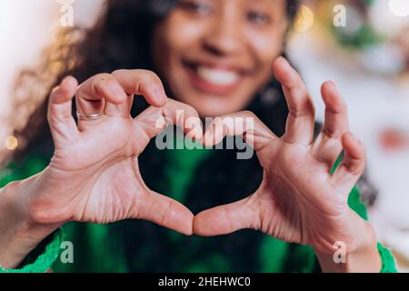 Brunette long-haired daughter and African American mother sit and pose showing hearts with fingers against Christmas decorations Stock Photo