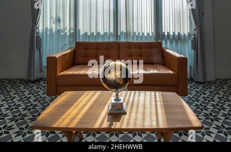 Chiang Rai,Thailand - Sep 06, 2020 : Replica globe on wooden coffee table front to brown leather sofa on background of white curtains and glass window Stock Photo