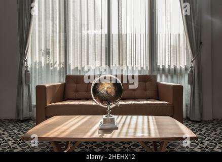 Chiang Rai,Thailand - Sep 06, 2020 : Replica globe on wooden coffee table front to brown leather sofa on background of white curtains and glass window Stock Photo