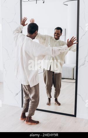 Positive young African-American guy in loose white shirt and trousers dances looking at large mirror reflection at home backside view Stock Photo