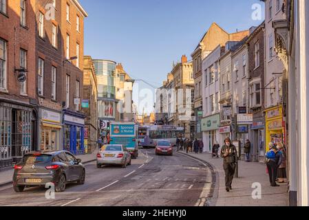 A city street with traffic and a queue of people wait at the bus stop.  Shops line the street  and people walk on the pavement. Stock Photo