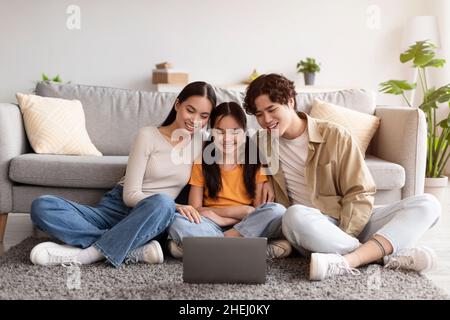 Satisfied millennial japanese mom, dad and teen daughter sitting on floor and watching video on laptop