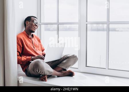Joyful African-American guy in stylish clothes laughs at video call via modern laptop sitting on large window sill at home Stock Photo