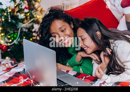 Curly-haired black mother and brunette daughter lie on bed near Christmas decorations and communicate via video call using laptop Stock Photo