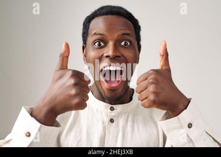 Portrait of emotional happy African-American guy in white shirt showing thumbs up posing on light grey background close view Stock Photo
