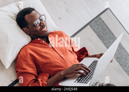 Cheerful African-American student with glasses in orange shirt works on laptop lying on floor near phone close upper view Stock Photo