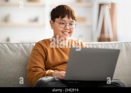 Cheerful chinese kid sitting on couch with laptop at home Stock Photo